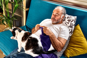 Bearded middle aged man sitting on sofa knits a violet wool scarf with the knitting needles of ball...