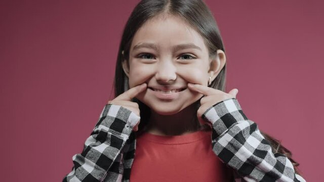 Little girl creates a smile with her fingers in pink studio, gives happy emotion. Cute kid smiling in the studio with pink background. High quality 4k footage