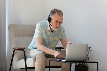 Focused mature old businessman in glasses wearing headset with mic, involved in communicating distantly by web camera video call using computer application, studying remotely on online courses.