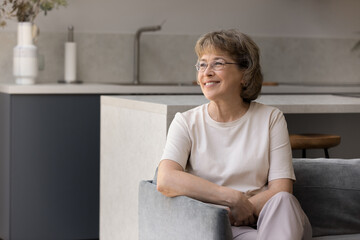 Thoughtful dreamy middle aged 60s retired woman in eyeglasses looking in distance, recollecting good memories, meditating, enjoying peaceful moment alone at home, resting on comfortable couch.