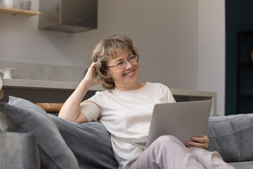 Addicted to modern technology dreamy smiling middle aged woman in eyeglasses using computer, thinking of getting pleasant news, enjoying shopping online or chatting with friends in social network.
