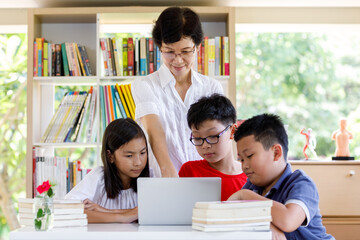 Fototapeta na wymiar A Group of Asian Student Kid Reading and looking Laptop on the table with female teacher in School library with Shelf of Books Background