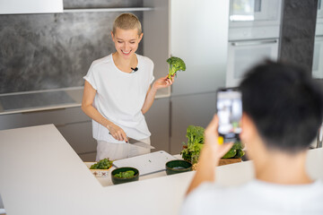 Man taking video on smartphone of caucasian woman cooking vegetarian salad on kitchen. Concept of video blogging. Idea of healthy and diet eating. Smiling girl with knife looking at mobile phone