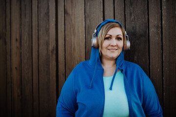 Happy overweight woman in sports clothes with headphones looking at camera against wooden wall