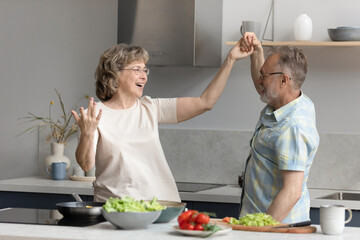Happy middle aged elderly retired married family couple dancing to music in modern kitchen,...