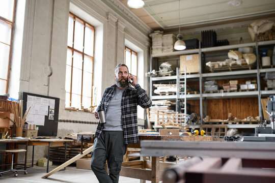 Happy mature male carpenter making phone call during coffee break indoors in carpentery workshop.