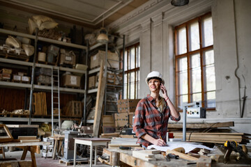 Fototapeta premium Female engineer holding blueprints and making phone call indoors in carpentry workshop.