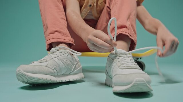 Boy tying shoelaces on sneakers sitting on a skateboard on blue background. Cute kid sitting in the studio with blue background. High quality 4k footage