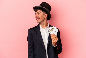 Young caucasian wizard man holding a magic cards isolated on white background looks aside smiling,...
