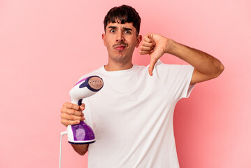Young mixed race man holding an iron isolated on pink background showing a dislike gesture, thumbs...