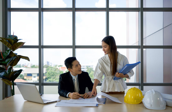 Asian project owner in suit and his assistant checking the construction drawing. Morning work atmosphere in a modern office.