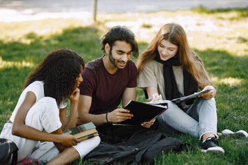 Three international students sitting on a grass in a park and holding a books