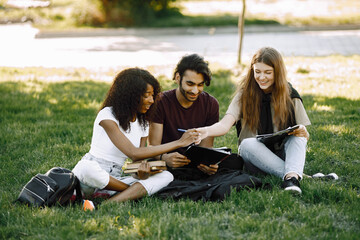 Three international students sitting on a grass in a park and holding a books