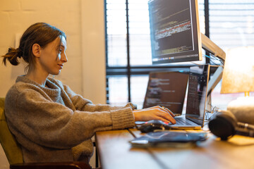 Young focused woman writing programming code, sitting at workplace with laptop and desktop computers in home office. Concept of freelance and remote work from home. Woman wearing domestic clothes