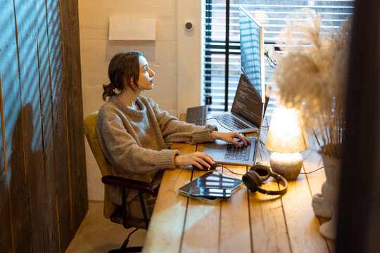 Young woman works on computers, sitting at workplace at cozy home office interior. Concept of freelance and remote work from home. Programmer writing code. Caucasian woman wearing domestic clothes.