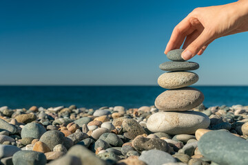Close-up of a woman's hand putting the last stone on top of pebbles on the seashore on a sunny day, copy space. Concept of balance, harmony