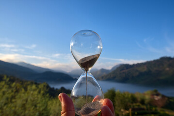 A hourglass (with falling sand) on a wooden wet table with sea of fog and mountain range silhouette...