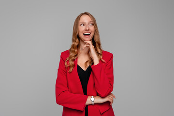 Smiling pretty woman has blonde hair looks aside with happy expression, plans something on coming weekends, standing in red blazer against gray background