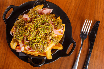 Porzione di patate fritte condite con mortadella, pesto e granella di pistacchio servite in un contenitore nero