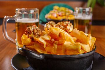Porzione di patate fritte condite con polpette di carne e cheddar fuso servite in un contenitore nero con sullo sfondo due boccali di birra e un altra porzione di patate fritte