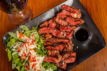 Tagliata di carne di Scottona cotta al sangue con contorno di rucola, pomodorini e scaglie di formaggio servita su un piatto nero e accompagnata da un calice di vino rosso