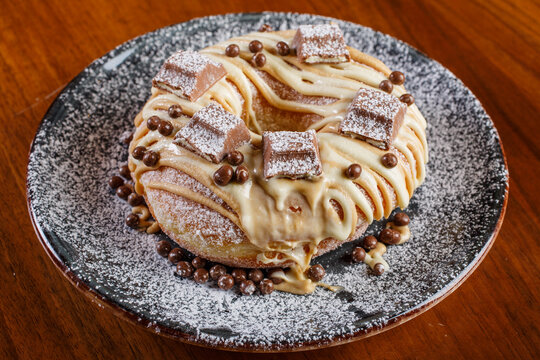 Graffa fritta guarnita con cioccolato bianco, praline di cioccolato al latte, barretta di cioccolata kinder e zucchero a velo servita su un piatto nero