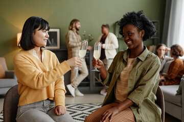 Young women toasting with flutes of champagne while sitting at party in the living room with other people in the background