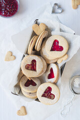Freshly baked Linzer cookies in a loaf tin set against a white background. Jam, cookie cutters, heart shaped biscuits included in frame. Flat lay style. Love, romance and valentines day food concept