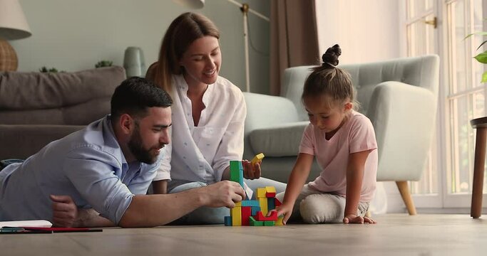 Caring mother father teach little daughter preschooler to create colorful buildings from brick constructor set on floor with underheating. Happy foster parents engage adopted girl in education game