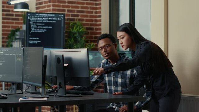Two software developers holding laptop with coding interface walking towards desk and sitting down talking about group project. Programmers team discussing algorithms pointing at computer screen.