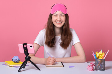 cheerful cute smiling brunette teen girl doing homework sit desk with smartphone fixed to tripod on white table happy studying at home online wearing a sleep mask distance quarantine education