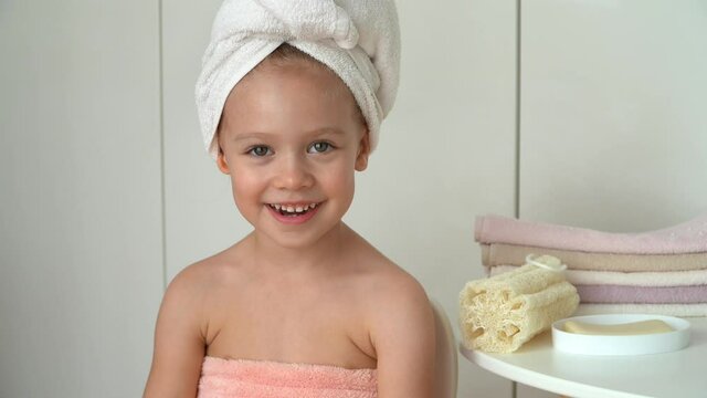 Portrait of little girl looks at camera and laughs in bathroom. Happy child after bathing with wrapped towel on head. Baby care.