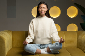 Time for yoga classes. Attractive young asian woman trains and sits in a yoga lotus position on the couch