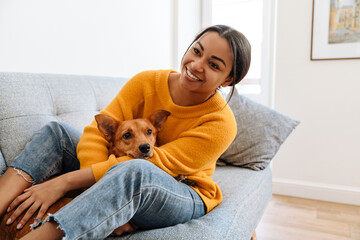 Young hispanic woman smiling while resting on couch with her dog