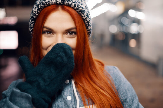 Outdoor night photo of young beautiful happy smiling girl enjoying festive decoration, in street of european city, wearing knitted beanie hat