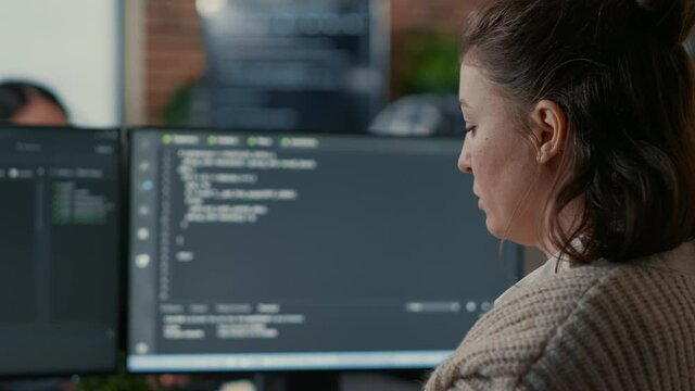 Over shoulder view of focused database designer writing code looking at multiple computer screens displaying algorithm. System engineer coding while colleagues doing teamwork in background.