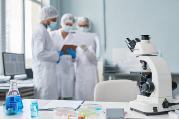 Close-up of workplace of scientist with microscope and lab glassware with liquid on the table with people in the background