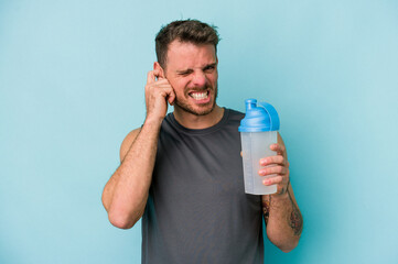 Young caucasian man drinking milkshake isolated on  blue background covering ears with hands.