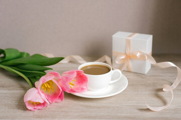 Fototapeta na wymiar Coffee with milk in a white porcelain cup and saucer, a bouquet of spring pink tulips and a gift in a white box with a satin ribbon. Mother's Day, Valentine's Day, Easter. The concept of spring