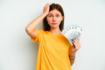 Young English woman holding banknotes isolated on blue background being shocked, she has remembered important meeting.