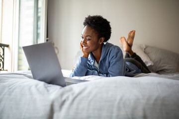 smiling young african american woman lying on bed looking at laptop computer