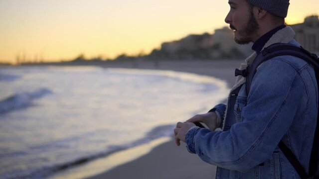 traveler of about 25 years making video with mobile phone on the beach during sunset. He wears a hat and a denim jacket. you can see the sunset in the background. He makes the video to have memories.