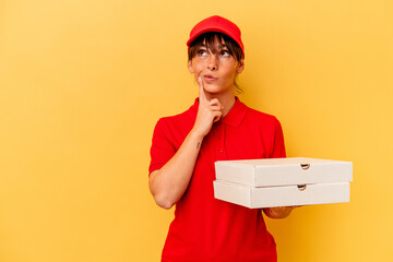 Young delivery woman holding pizzas isolated on yellow background looking sideways with doubtful and skeptical expression.