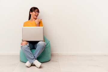 Young Argentinian woman working with pc sitting on a puff isolated on white background points with thumb finger away, laughing and carefree.