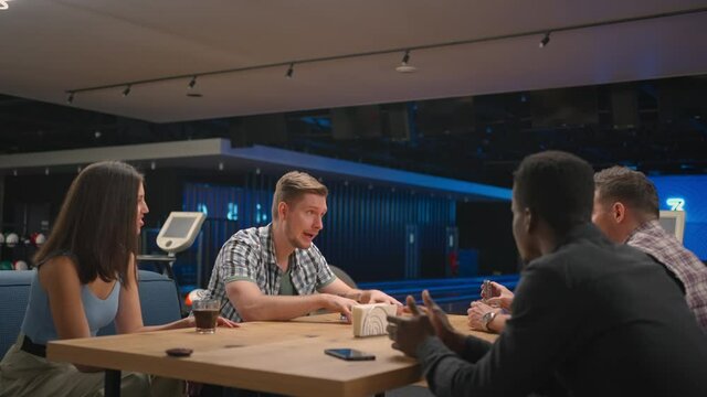 A multi ethnic group of friends in a bowling club communicate at the table and rejoice in the victory of a friend