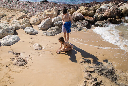 young woman man on the beach. A girl in love draw a heart on the sand, sitting on the seashore. Valentine's Day on vacation, first love, happy relationship, summer vacation, sunbathing