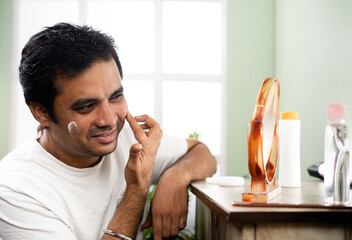 Handsome young man applying anti-aging or skin moisture cream on face by looking mirror at home -...