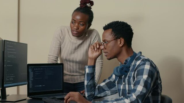Database developer removing glasses while explaining algorithm to colleague coder pointing at running code on multiple computer screens. System engineers collaborating on development group project.