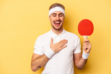 Young caucasian man holding ping pong rackets isolated on yellow background laughs out loudly keeping hand on chest.