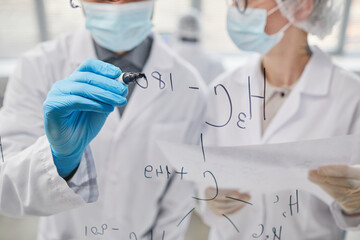 Close-up of two scientists writing chemical formulas on glass board and discussing them in team
